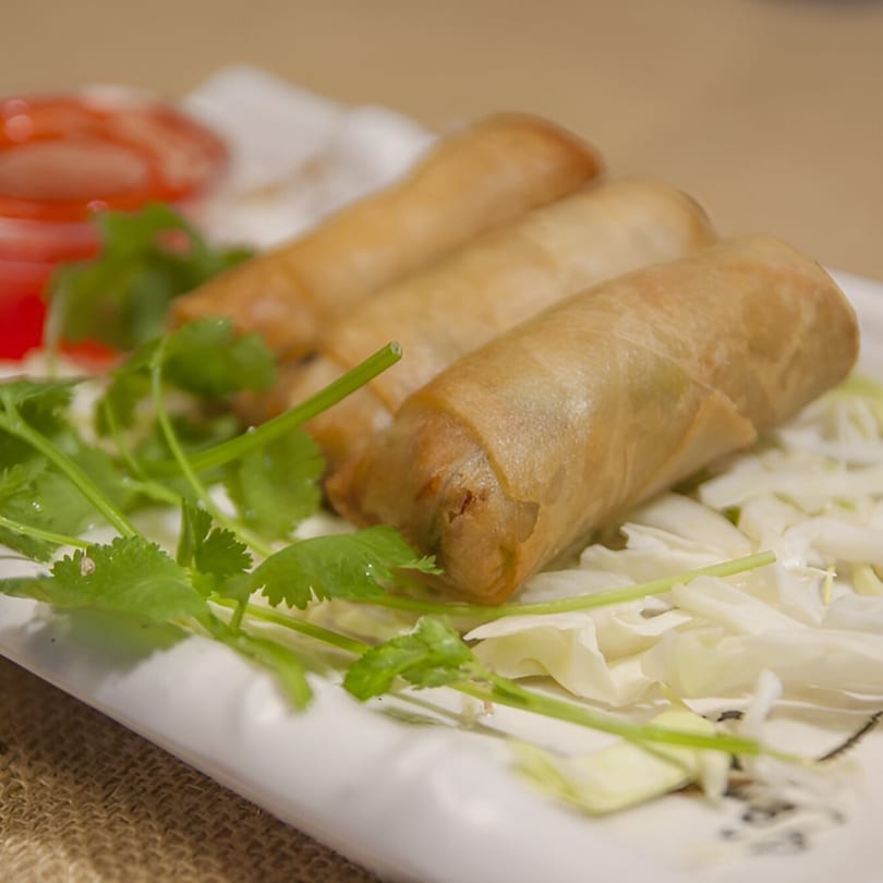 Three golden-brown vegetarian spring rolls on a white plate with shredded cabbage, cilantro, and red sauce.